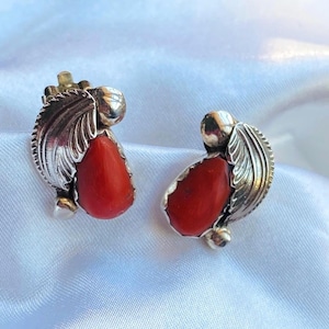May include: A pair of silver-toned stud earrings featuring a leaf design and a teardrop-shaped, vibrant red coral stone. The earrings are set against a white background.