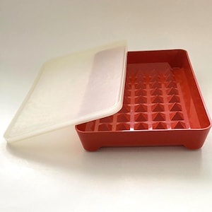 May include: A red plastic tray with a textured interior and a translucent lid. The tray has a grid of pyramid-shaped bumps. The lid is slightly ajar, revealing the tray's interior. The tray is rectangular with rounded corners.