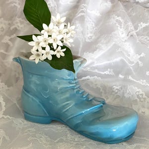 May include: A light blue ceramic boot-shaped vase with white flowers and green leaves. The vase has a glossy finish and is decorated with laces. The flowers are clustered together, creating a delicate floral arrangement.