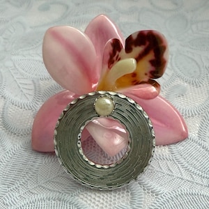 May include: A circular silver-toned brooch featuring a textured, grooved surface and a single central cream-colored faux pearl accent. The brooch has a scalloped outer edge and a smooth inner rim, presenting a metallic finish with a polished appearance.