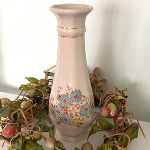 May include: A cream-colored ceramic vase with a flared top and a hexagonal base. The vase features a floral design with blue and pink flowers. The vase is decorated with brown accents and is surrounded by a garland of green leaves and berries.