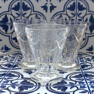 May include: A set of three clear glass tumblers with a footed base and a decorative embossed wave pattern around the upper rim. Each glass features a vertical ribbed design at the bottom section, providing a textured grip and a classic aesthetic.