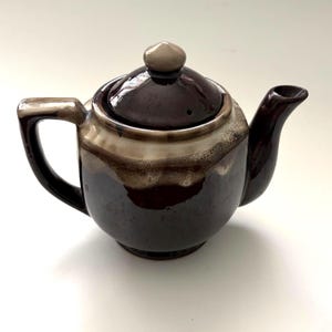 May include: A brown ceramic teapot with a lid and spout. The teapot has a rounded body and a handle. The lid is topped with a small, rounded knob. The teapot has a two-tone glaze, with a lighter color at the top and a darker brown at the bottom.