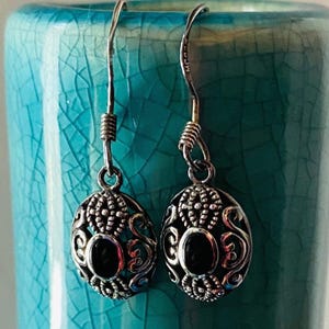 May include: Silver filigree earrings with an oval black stone center. The earrings have a decorative design and are suspended from a silver hook. The background is a turquoise blue ceramic surface.