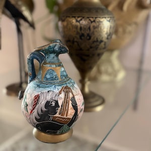May include: A small ceramic pitcher featuring a blue and white marbled finish with gold accents. The front displays a black silhouette illustration of a figure playing a lyre, accented with red and green details. It has a handle and a gold base.