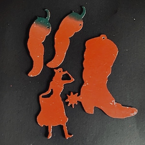 May include: Four orange metal cutouts: two chili peppers with green stems, a cowboy boot, and a figure of a person wearing a hat and a star-shaped spur. The cutouts are on a black background.