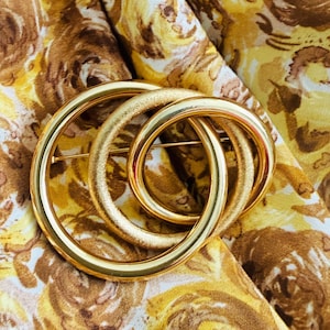 May include: A gold-toned brooch featuring three interlocking circular rings. Two of the rings have a smooth, polished finish, while the central ring displays a textured, matte surface. The brooch is positioned against a yellow and brown floral fabric.