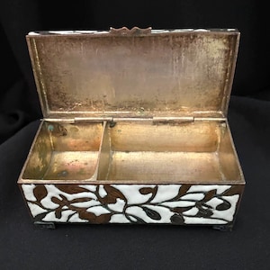 May include: An open, antique jewelry box with a hinged lid. The box is rectangular with a white and brown floral design on the exterior. The interior is gold-toned with two compartments. The box is resting on small feet.