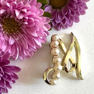 May include: A gold-toned letter "A" brooch with a pearl accent. The brooch features a cursive design with a row of six faux pearls. The background includes vibrant pink flowers.