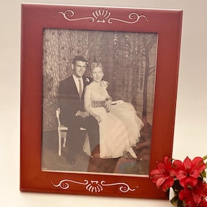 May include: A framed black and white photograph of a couple, set in a red wooden frame with decorative white accents. The photo shows a man in a suit and a woman in a dress. Red flowers are in the foreground.