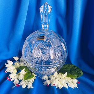 May include: A clear, spherical lead crystal candy dish featuring intricate starburst and cross-hatch cut patterns. The lid is topped with a faceted, diamond-shaped finial. A small rectangular gold and blue sticker is affixed to the upper surface.