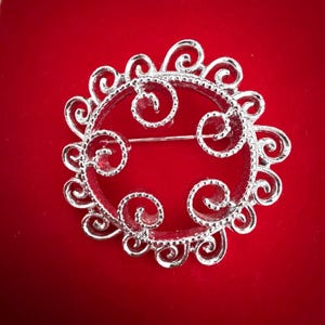 May include: A circular silver-toned brooch featuring an intricate openwork design with multiple swirling scroll patterns along the outer edge and three smaller spirals extending inward from the inner rim, finished with a polished metallic surface.