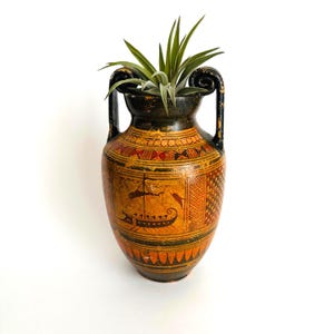 May include: A ceramic amphora vase featuring black and orange geometric patterns and a painted scene of a ship with a sail. The vase has two curved handles and contains a green air plant with pointed leaves extending from the top opening.