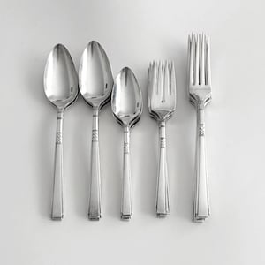 May include: A set of five silver-toned metal utensils including three spoons of varying sizes and two forks. Each piece features a smooth, polished finish with a decorative geometric band detail on the handle and a straight, tapered stem design.