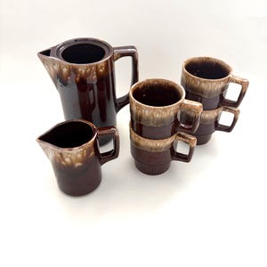 May include: A ceramic coffee set featuring a tall pitcher, a smaller creamer pitcher, and four stackable mugs. Each piece has a dark brown base with a lighter beige drip glaze pattern around the top rim, creating a distinct two-tone appearance.