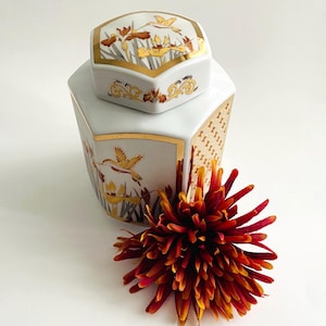 May include: A white ceramic decorative box with a lid, featuring gold trim and floral and bird designs. The box has a hexagonal shape and is accented with gold patterns. A red and yellow flower is in the foreground.