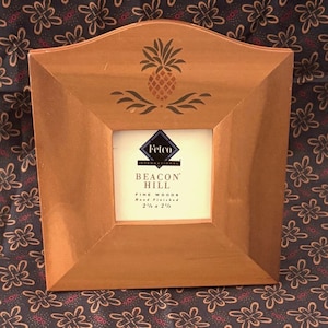 May include: A light brown wooden picture frame with a curved top edge and a dark brown pineapple stencil design. The center insert features the text Fetco International Beacon Hill Fine Woods Hand Finished 2 1/2 x 2 1/2 inches.