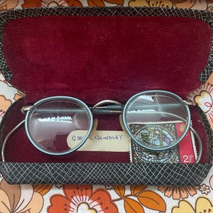 Peut inclure: Étui à lunettes vintage ouvert avec des lunettes rondes à monture bleu clair. L'étui est noir avec un motif en losange et un intérieur en velours rouge. Comprend une petite carte avec le nom "Cary Grindley" et un timbre-poste.