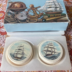 Könnte beinhalten: Ein Set aus zwei runden weißen Untersetzern mit einer nautischen Illustration eines Segelschiffs auf dem Ozean. Die Untersetzer befinden sich in einer hellblauen Schachtel, die mit einem Globus, einem Sextanten, einem Fernrohr und einem Schiff verziert ist.