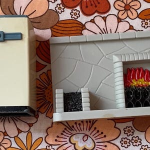 Puede incluir: Un pequeño refrigerador crema con un asa azul y una base negra está al lado de una chimenea gris. La chimenea tiene un patrón de piedra, un fuego rojo y amarillo y una rejilla negra.
