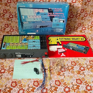 May include: A Science Fair 200-in-one electronic project lab kit featuring a grey console with a breadboard, a blue storage box, a red instruction manual, various coloured wires, and a small bag of components. The console includes knobs, a meter, and a speaker.
