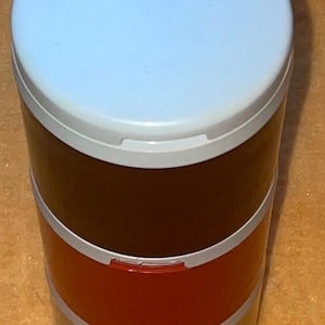 Könnte beinhalten: Ein mehrstöckiger Lebensmittelbehälter mit einem hellblauen Deckel, einem dunkelbraunen Mittelteil, einem roten Abschnitt und einem grauen Sockel. Der Behälter hat eine zylindrische Form und ist zum Transport von Lebensmitteln bestimmt.