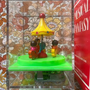 May include: A musical carousel toy enclosed in a clear acrylic case, featuring a yellow canopy with blue dots, a bright green base, and small figures riding colorful animals. The red packaging box displays the text Musical Fantasy in white lettering.