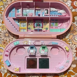 Op de afbeelding: Een vintage roze Polly Pocket compact speelgoed, opengeklapt om een miniatuurhuis met kamers, meubels en een trap te onthullen. Het onderste gedeelte heeft make-up-achtige compartimenten en kleine accessoires. Het speelgoed staat tegen een bloemenachtergrond.