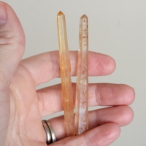 May include: Two orange-toned quartz crystal points are held in a hand. The crystals have a translucent appearance with variations in color and texture. The crystals are pointed at the top and have a natural, raw aesthetic. The background is a neutral, light color.