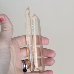 May include: Two clear quartz crystal points with a light orange hue are held in a hand. The crystals have a natural, rough texture on one side and a smooth, polished appearance on the other. The crystals are a light, translucent color.