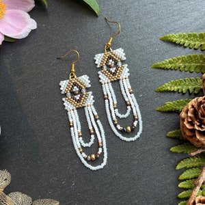 May include: White and gold beaded earrings with a geometric pattern. The earrings are hanging from a dark surface.
