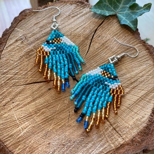 May include: A pair of beaded earrings with a turquoise, white, and gold color scheme. The earrings are teardrop shaped and have a fringe design.