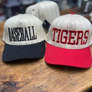 May include: Baseball caps with cream-colored tops and black or red brims. One cap has "BASEBALL" embroidered in black, and the other has "TIGERS" in red. Both caps have vertical pinstripes.