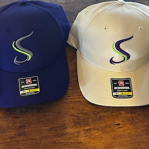 May include: Four baseball caps with a stylized "S" logo embroidered on the front. The caps are in gray, white, blue, and dark gray. The logo is purple and green.