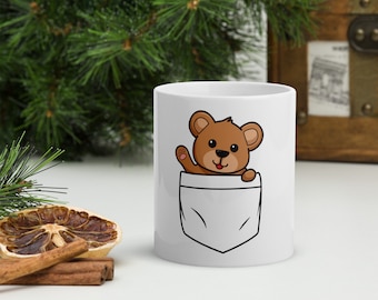 Op de afbeelding: Witte keramische mok met een bruine teddybeer die uit een witte zak op de voorkant kijkt. De beer glimlacht en zwaait.