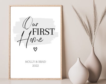 May include: A framed print with the words "Our First Home" in black script and block letters, with a small heart symbol. The names "Molly & Brad" and the year "2022" are below. The frame is light brown.