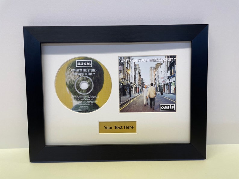 May include: A framed CD and album cover of Oasis's "What's the Story (Morning Glory)?" album. The CD is in a black frame with a white mount. The album cover is a photo of a street in London. The text "Your Text Here" is on a gold plaque below the album cover.