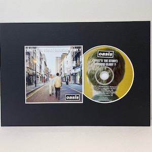 May include: A black framed CD and album set featuring the Oasis album "What's the Story) Morning Glory?". The album cover shows three men walking down a street in a city. The CD features a close-up of a man's head with the album title.