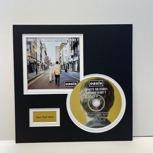 May include: Framed Oasis album art and CD for "(What's the Story) Morning Glory?". The framed image shows a street scene with two people. The CD is displayed alongside the frame. A small gold plaque is below the framed image.