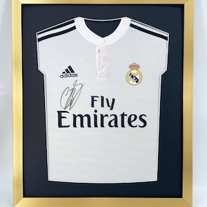 May include: A white soccer jersey with black stripes and the Adidas logo. The jersey has the Real Madrid crest and the words "Fly Emirates" printed on the front. The jersey is framed in a gold frame with a black mat.