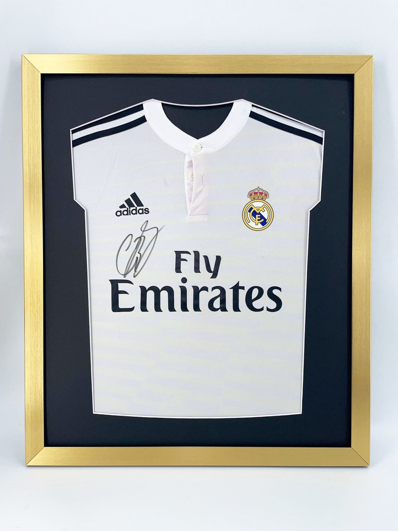 May include: A white soccer jersey with black stripes and the Adidas logo. The jersey has the Real Madrid crest and the words "Fly Emirates" printed on the front. The jersey is framed in a gold frame with a black mat.