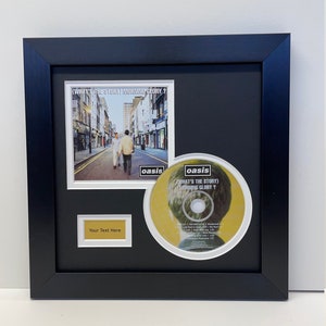 May include: A framed CD and album cover set featuring the Oasis album "(What's the Story) Morning Glory?" The album cover features a black and white photo of a street scene with two people walking. The CD is yellow with the band's name and album title printed in black. The frame is black with a gold plaque that says "Your Text Here".