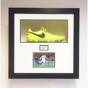May include: Framed display featuring a neon yellow football boot with a black Nike swoosh. Below the boot is a small photo of a footballer in action. The frame is black with white mount.