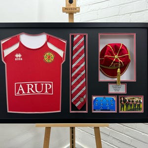 May include: A framed display of a red and white jersey with the word "ARUP" on the front, a red and white striped tie, a red cap with gold trim, and a team photo. The jersey has the word "errea" on the front.