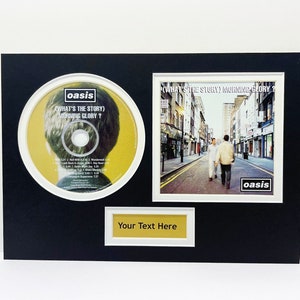 May include: A black framed display featuring a CD and album cover of Oasis's "What's the Story (Morning Glory?)". The CD is yellow with a picture of a man's head. The album cover is a photo of three men walking down a street. The text "Your Text Here" is on a gold label.