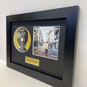 May include: A framed black and white photograph of a street scene with two people walking in the middle of the street. The photo is framed with a black frame and a gold plaque with the text "Your Text Here". The photo is accompanied by a CD with a yellow background and the text "oasis WHAT'S THE STORY) MORNING GLORY?" on it.