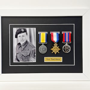 K&ouml;nnte beinhalten: Ein Schwarzwei&szlig;foto eines Mannes in Milit&auml;runiform mit drei ausgestellten Medaillen in einem wei&szlig;en Rahmen mit schwarzem Passepartout. Die Medaillen befinden sich auf einem schwarzen Hintergrund mit einer goldenen Inschrift, die "Your Text Here" lautet.