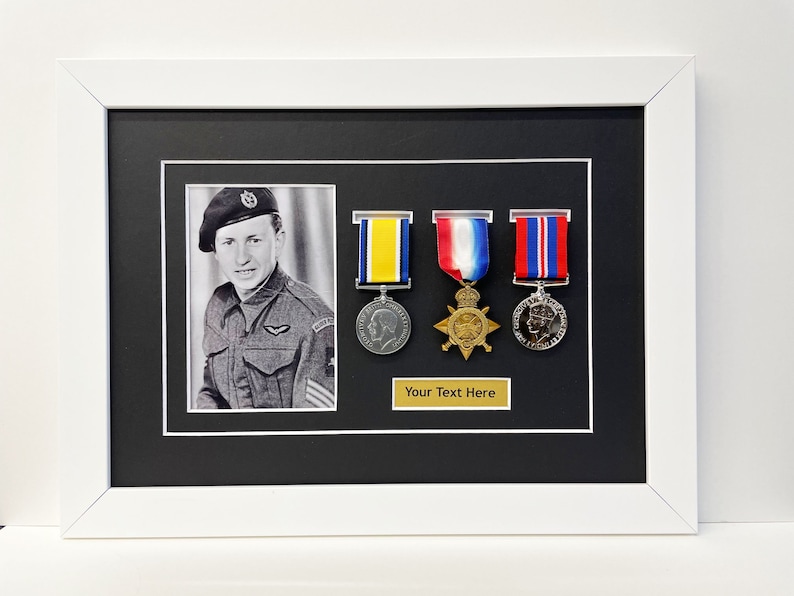 K&ouml;nnte beinhalten: Ein Schwarzwei&szlig;foto eines Mannes in Milit&auml;runiform mit drei ausgestellten Medaillen in einem wei&szlig;en Rahmen mit schwarzem Passepartout. Die Medaillen befinden sich auf einem schwarzen Hintergrund mit einer goldenen Inschrift, die "Your Text Here" lautet.