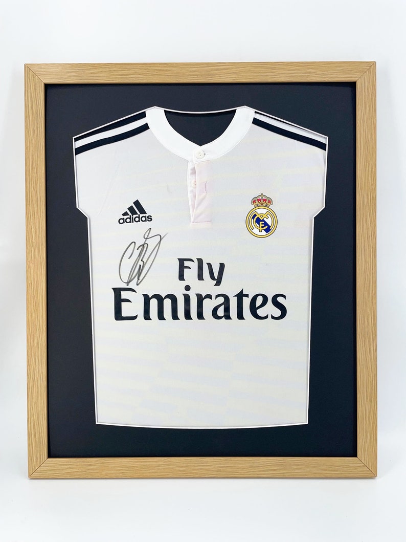 May include: A white soccer jersey with black trim and the Adidas logo. The jersey has the Real Madrid crest and the text "Fly Emirates" on the front. The jersey is signed with a black marker.