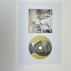 May include: A framed CD and album cover for Oasis's "(What's the Story) Morning Glory?" album. The album cover shows a street scene with two figures. The CD is gold with the album title and artist name.